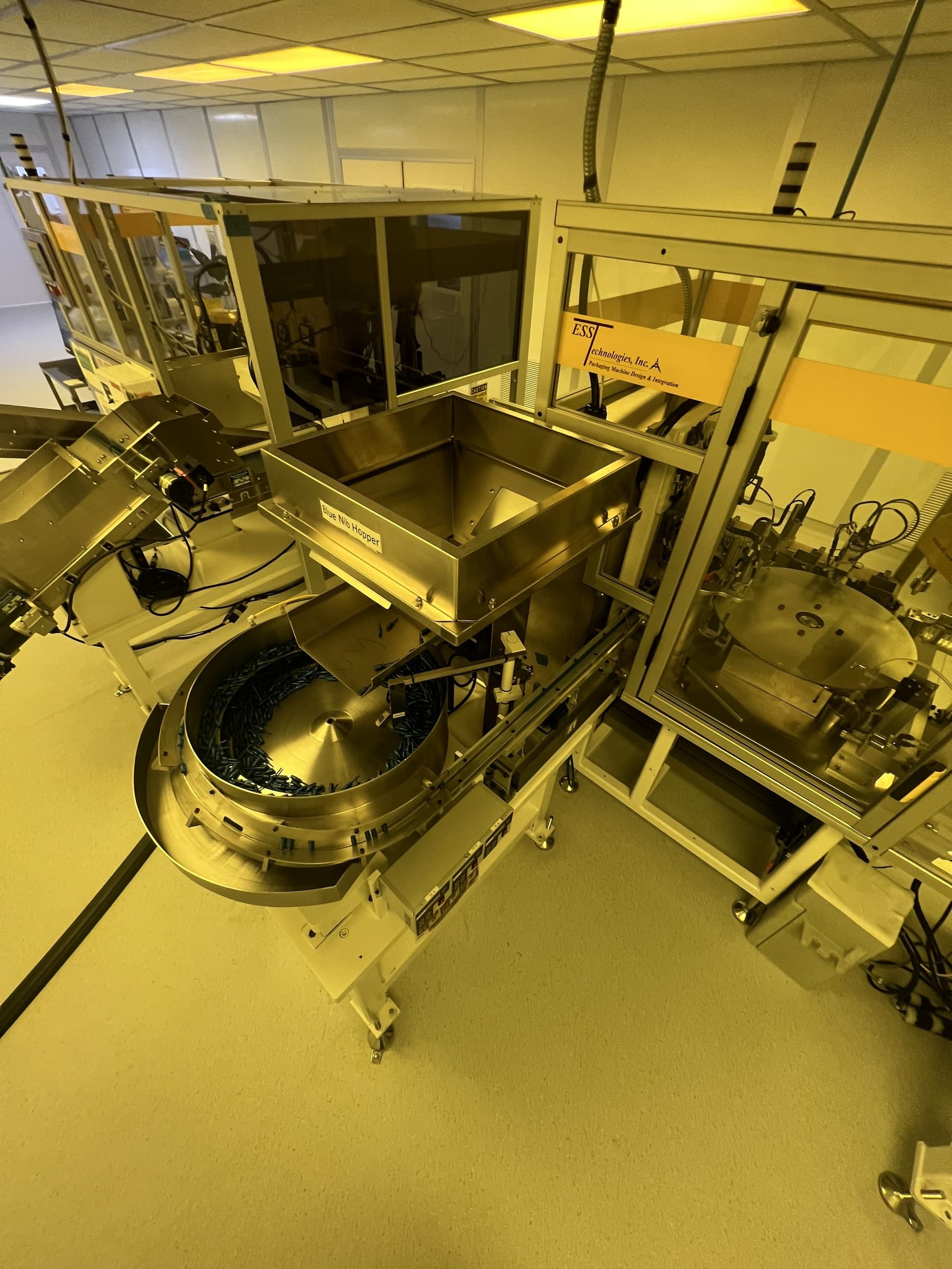 Hopper and circular feeder beside enclosed production machinery under yellow lighting.